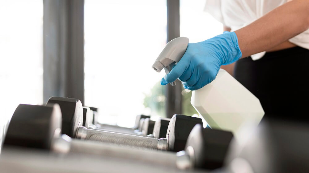 A cleaner in Fort Worth, TX, meticulously disinfecting gym equipment. Wearing gloves and using a spray bottle, the individual is focused on sanitizing a treadmill to ensure a hygienic environment for gym users.